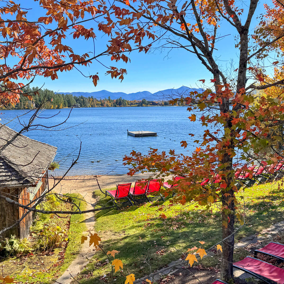 blue lake in Lake Placid surrounded by orange and red trees with red chairs lined up in front of lake and blue skies above.