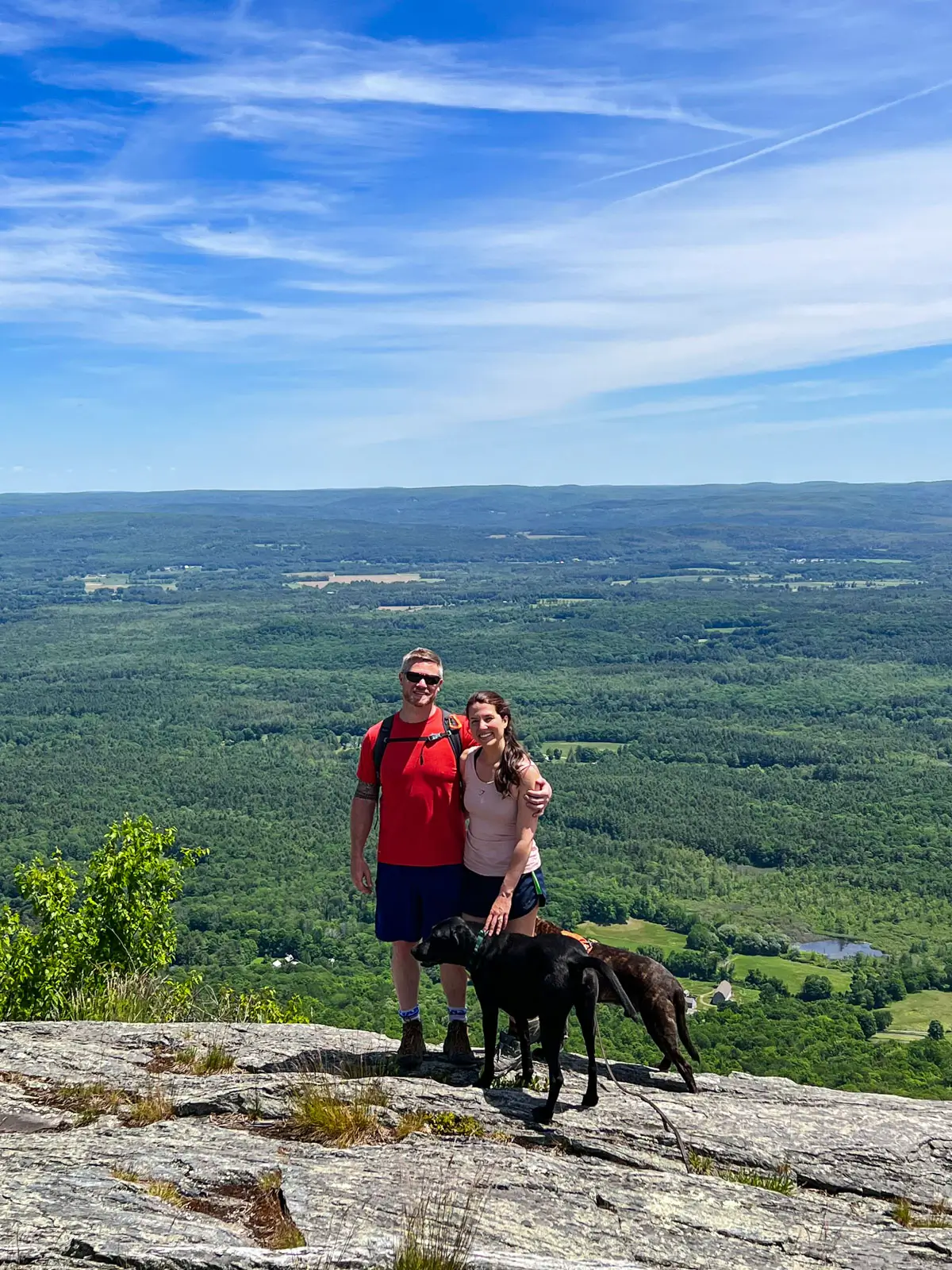 woman in pink tank top and man in red t-shirt in summer at the top of mount race with green hills and farmland in distance and blue sky above.