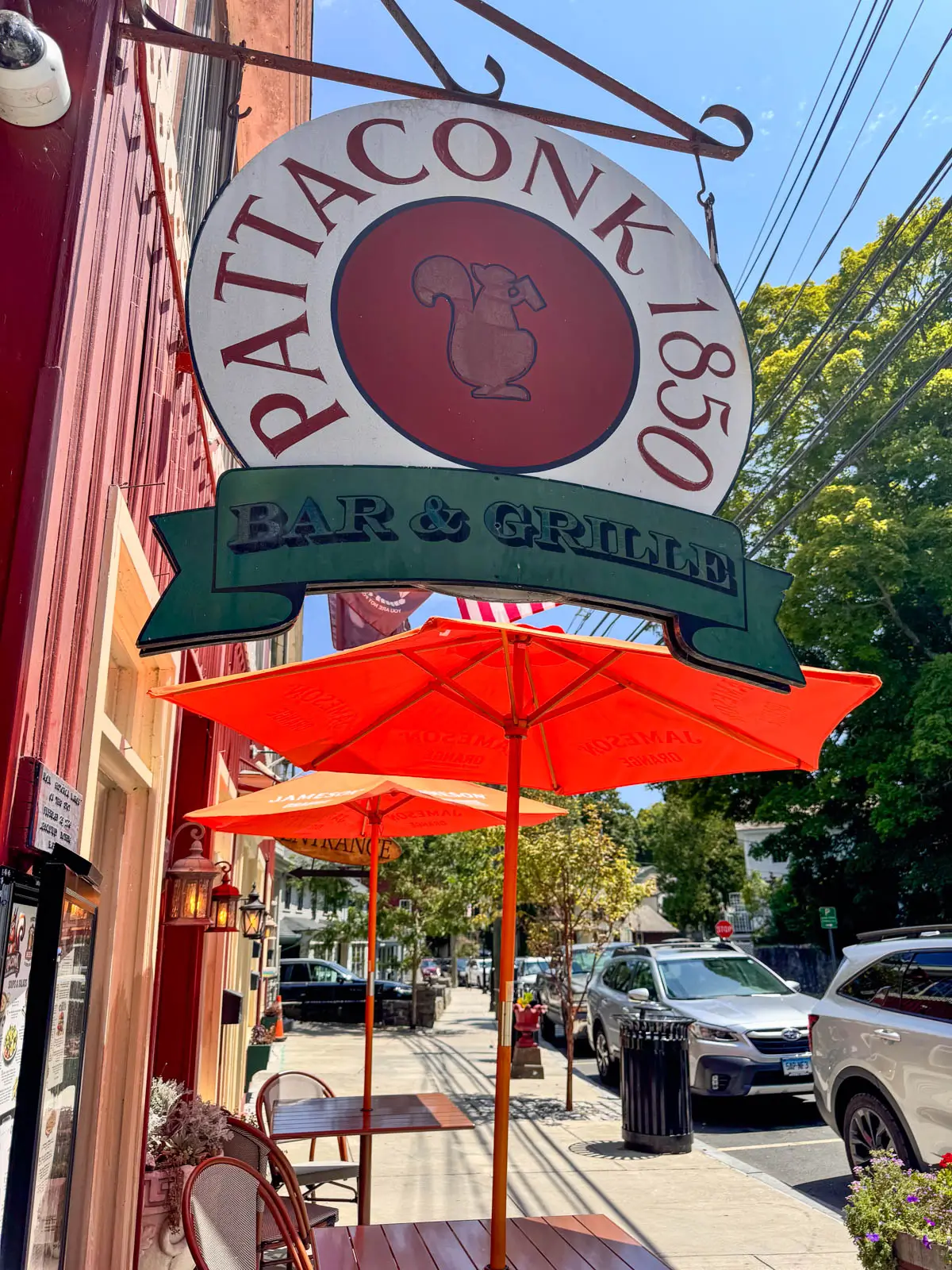 outside of a restaurant in chester with round sign hanging from side of building with "pattaconk 1850" written on the sign and two table with orange umbrellas out front.