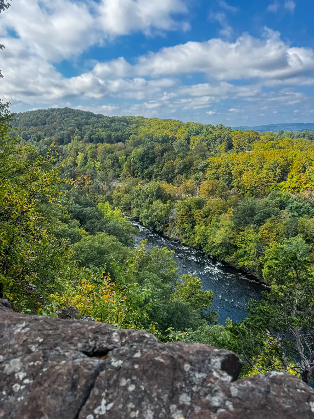 View from top of hike overlooking blue river below and bright green trees all around and blue sky above with white fluffly clouds at hatchet hill in tariffville, connecticut.