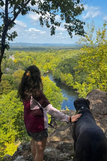 woman standing on top of cliff with big brown dog overlooking blue river below and bright green trees all around.