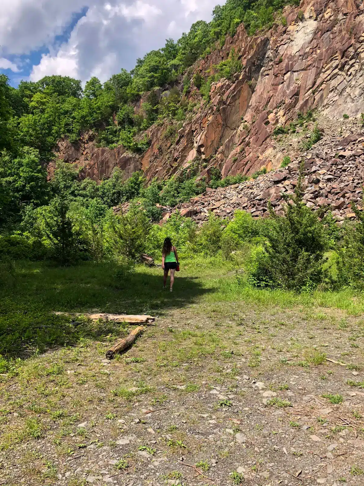 woman in bright green tank top and blue shorts walking at the base of a quarry with patched of green trees at sleeping giant state park in CT.
