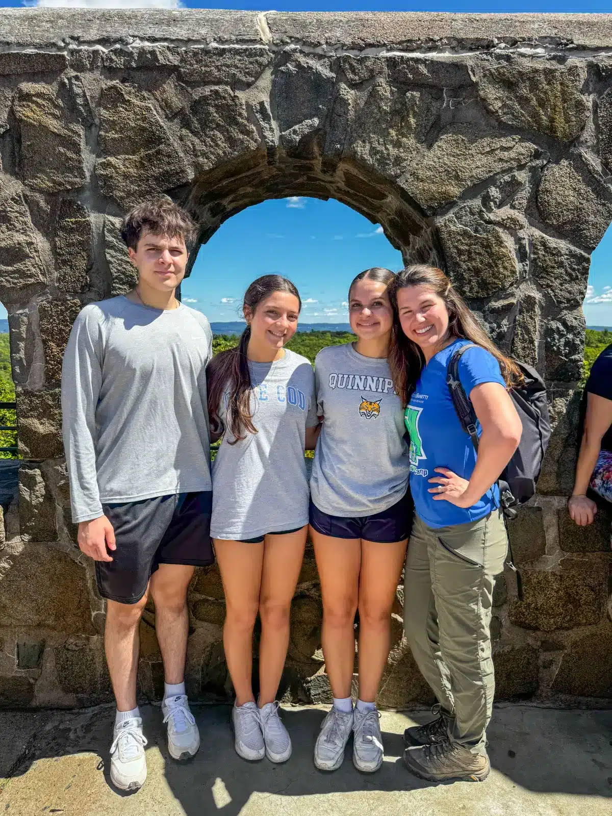 woman in blue short sleeve shirt and two teenage girls next to her in gray T shirts and tall teenage boy in gray shirt at top of stone tower in hamden.