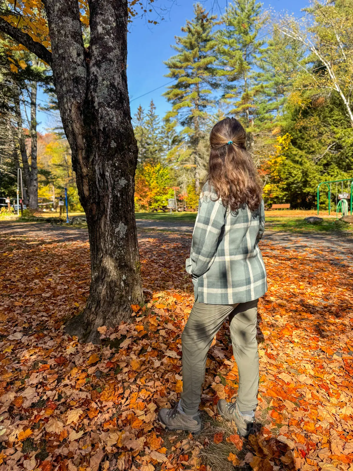 back of brunette woman on a fall day in vermont with orange leaves on the ground wearing an oversized green flannel jacket with hands in the flannel's pockets.