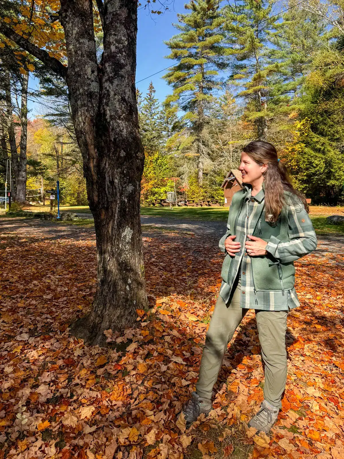brunette woman on a fall day in vermont with orange leaves on the ground wearing an oversized green flannel jacket with hands in the flannel's pockets.