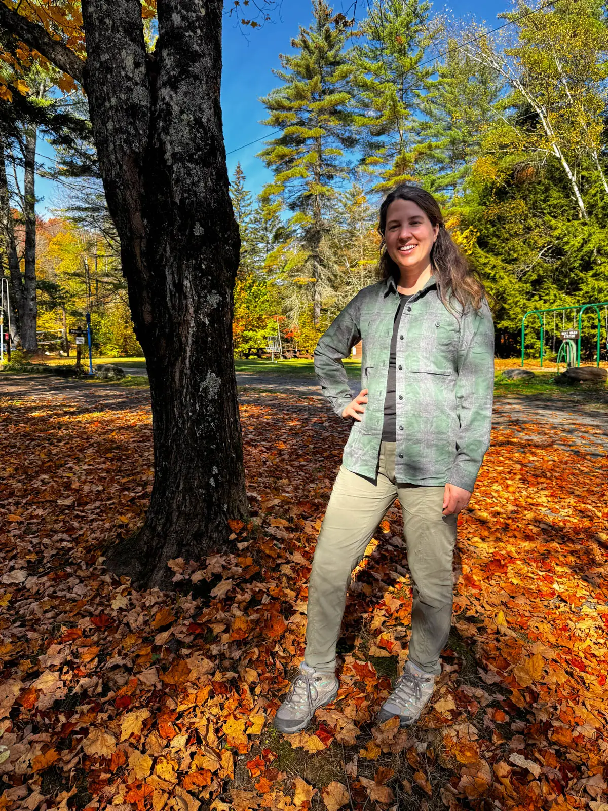 brunette woman smiling at the camera on a fall day in vermont wearing an a gray and green light flannel shirt with one hand on her hip.