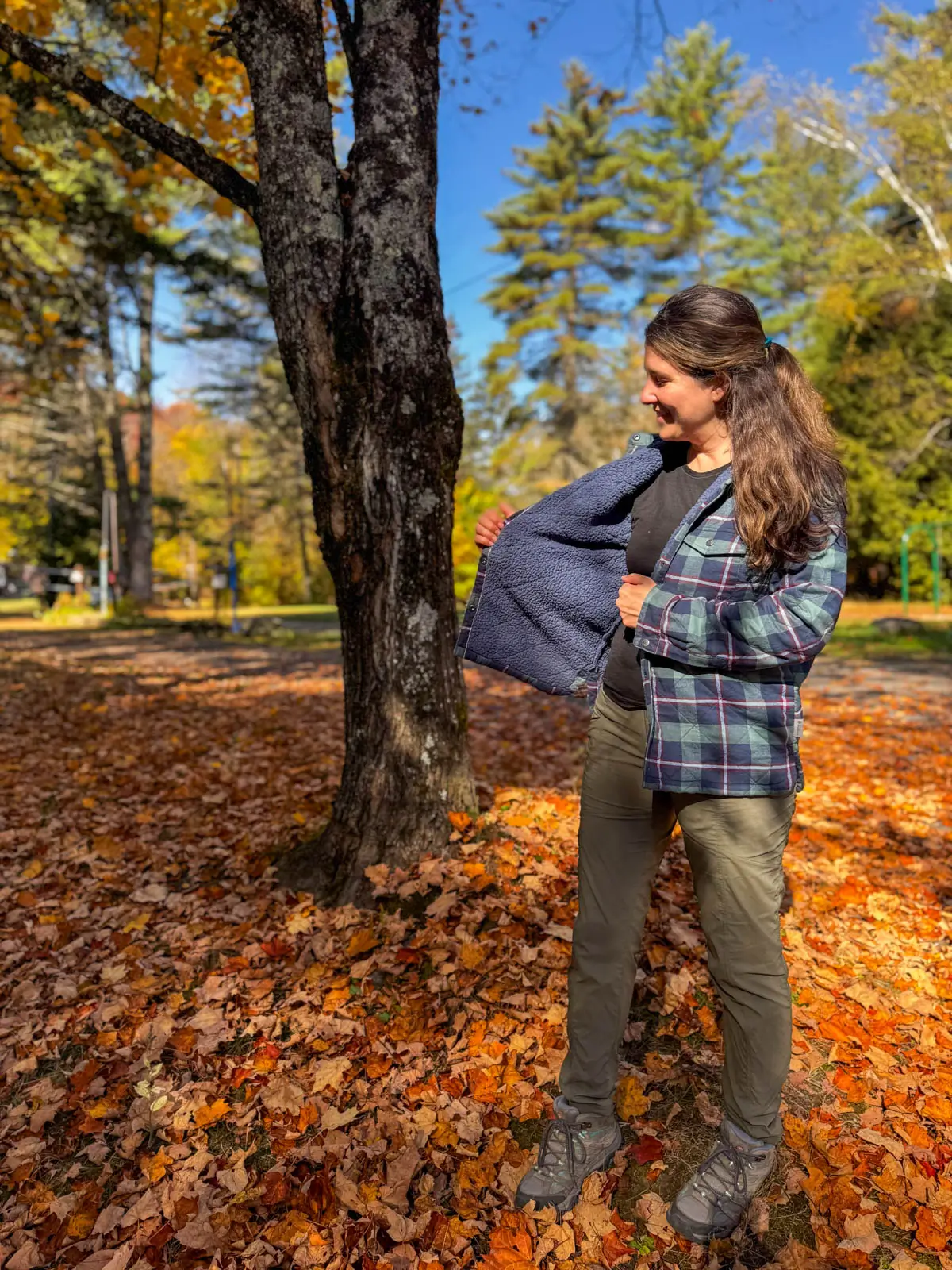 brunette woman smiling at camera standing in orange leaf pile in vermont wearing a navy blue and green checkered flannel with fleece lining on a sunny day.