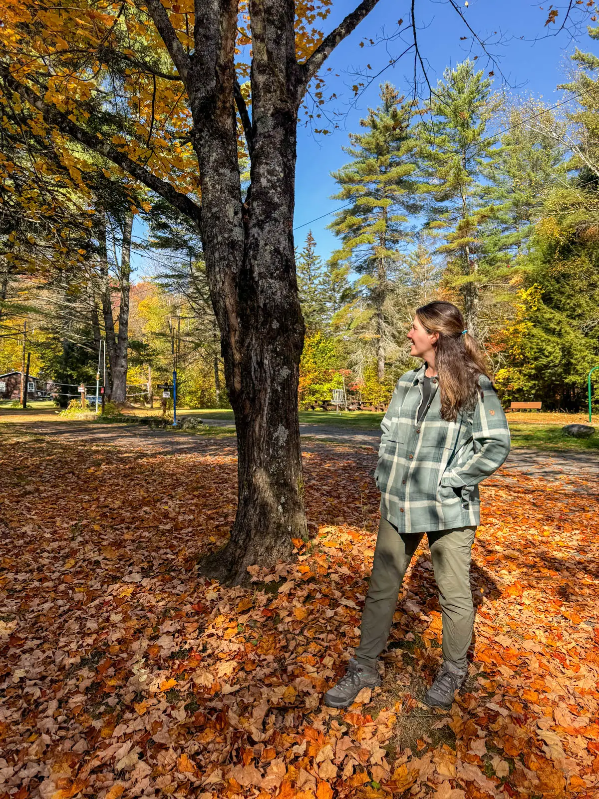 brunette woman smiling and looking off to the side on a fall day in vermont wearing an oversized army green flannel jacket with hands in the flannel's pockets.