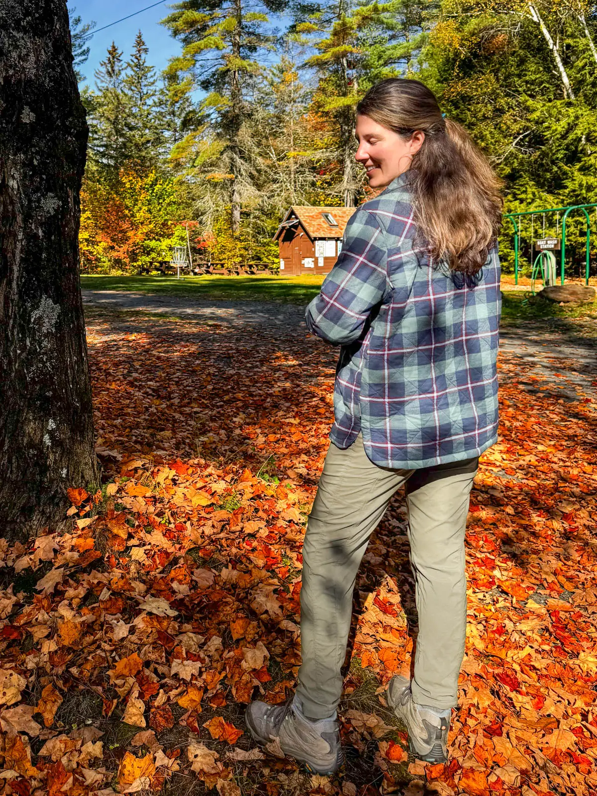 brunette woman smiling at camera standing in orange leaf pile in vermont wearing a navy blue and green checkered flannel with fleece lining on a sunny day and holding open one side of the flannel so you can see the navy fleece lining underneath.