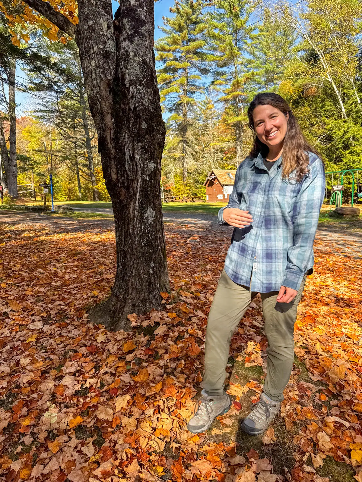 brunette woman standing on orange leaf pile smiling at camera in a light blue checkered flannel shirt with hiking boots and green pants on.