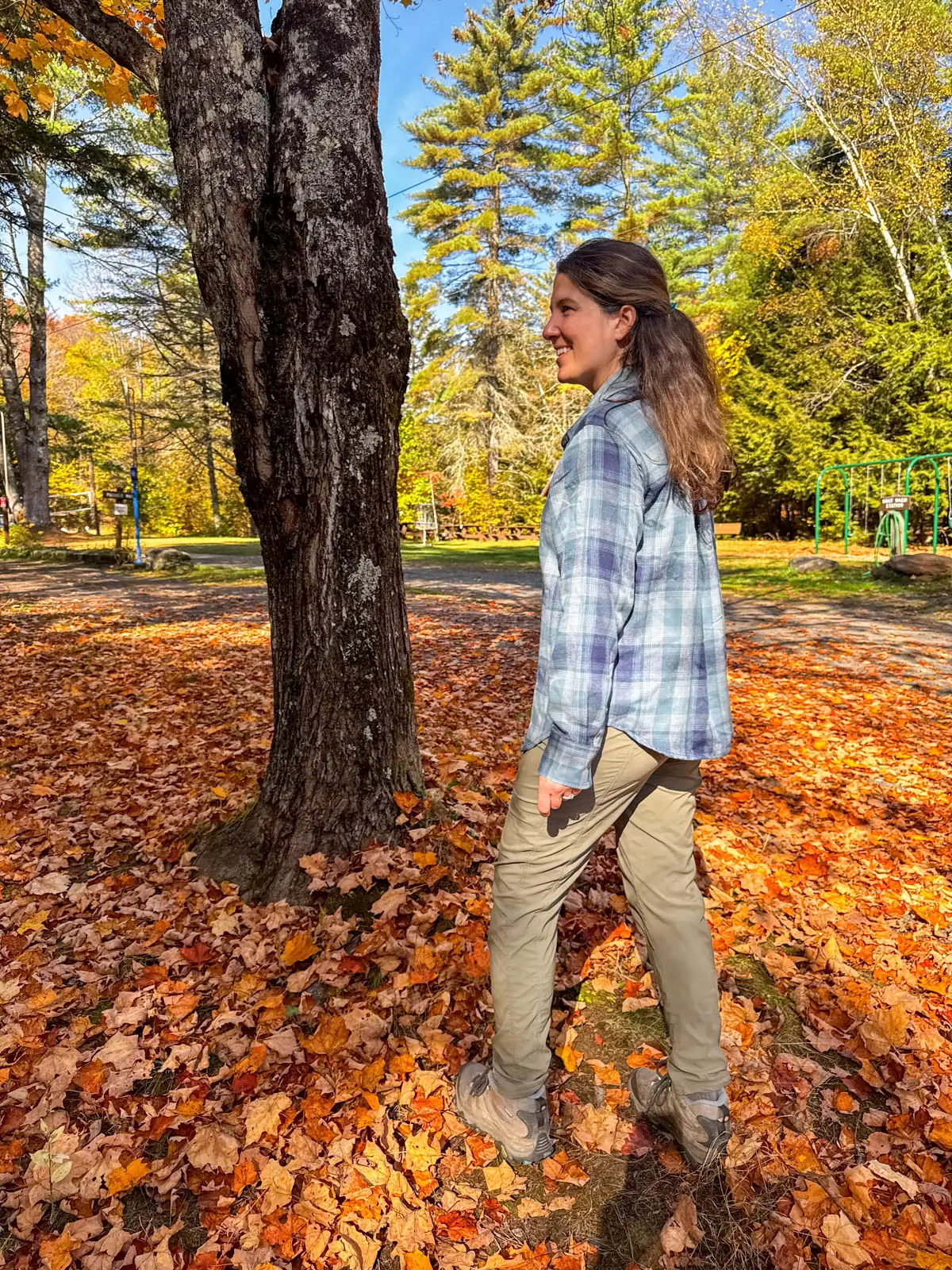 brunette woman standing on orange leaf pile smiling over her shoulder in a light blue checkered flannel shirt with hiking boots and green pants on.