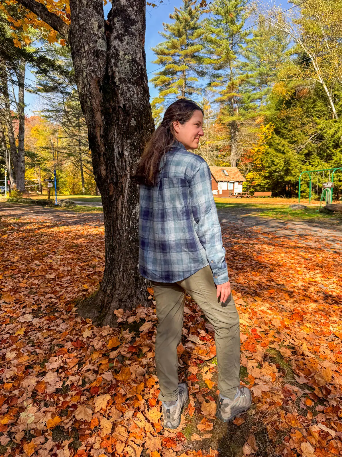 brunette woman standing on orange leaf pile smiling over her shoulder in a light blue checkered flannel shirt with hiking boots and green pants on.