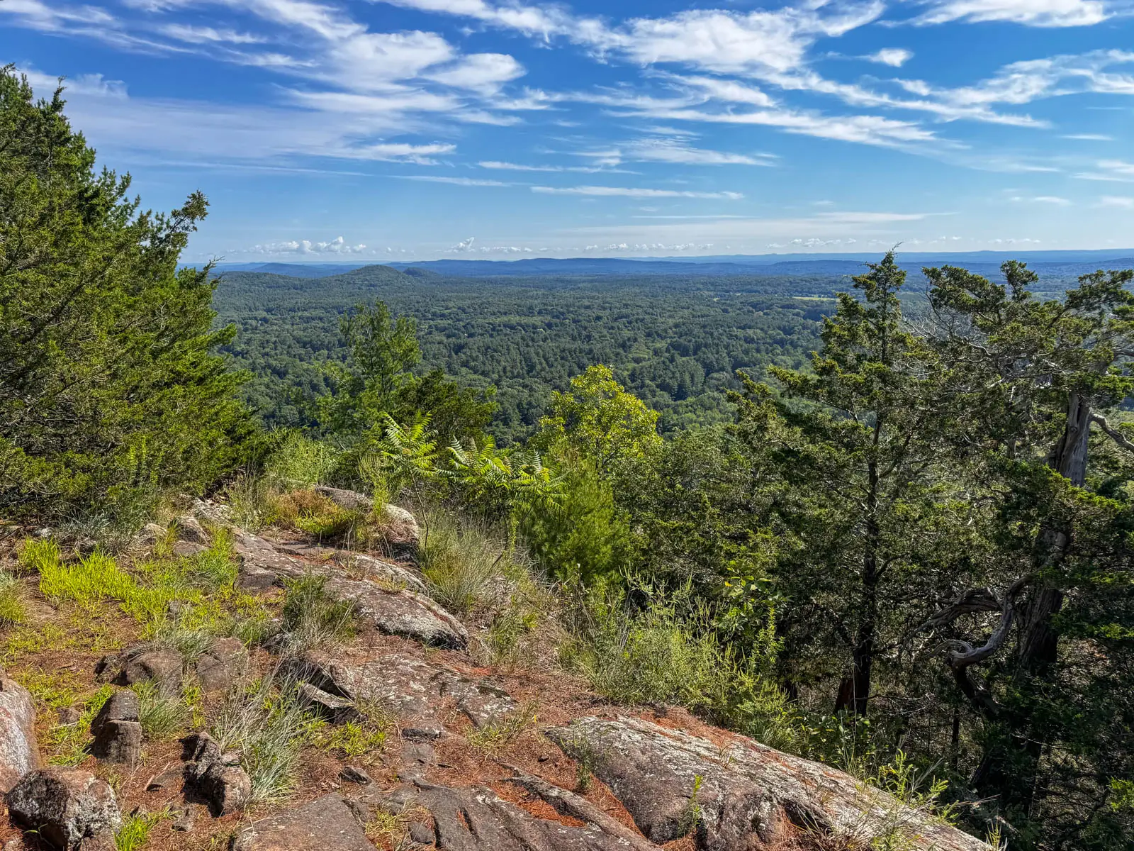 view from top of small mountain with green landscape in distance and blue sky with small fluffy white clouds above.