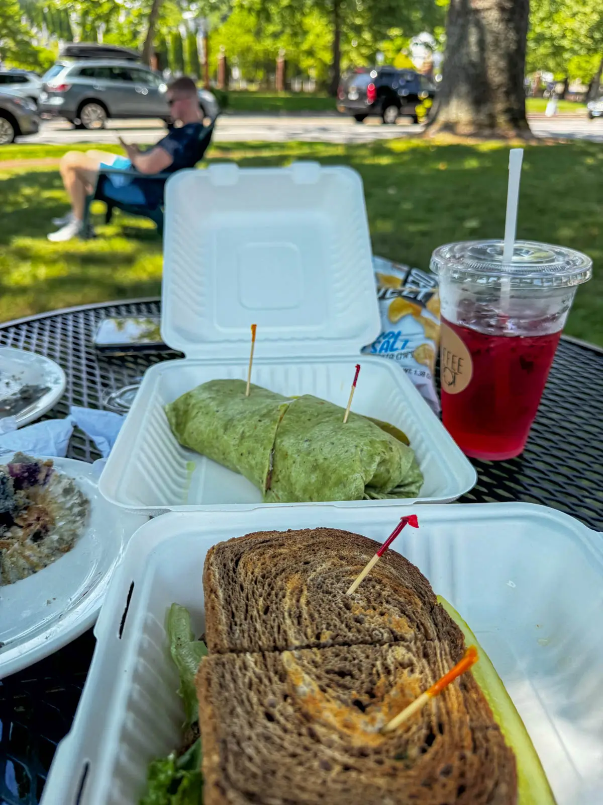 to go containers filled with a spinach wrap in one and a rye bread sandwich in the other with a red iced tea on the table.