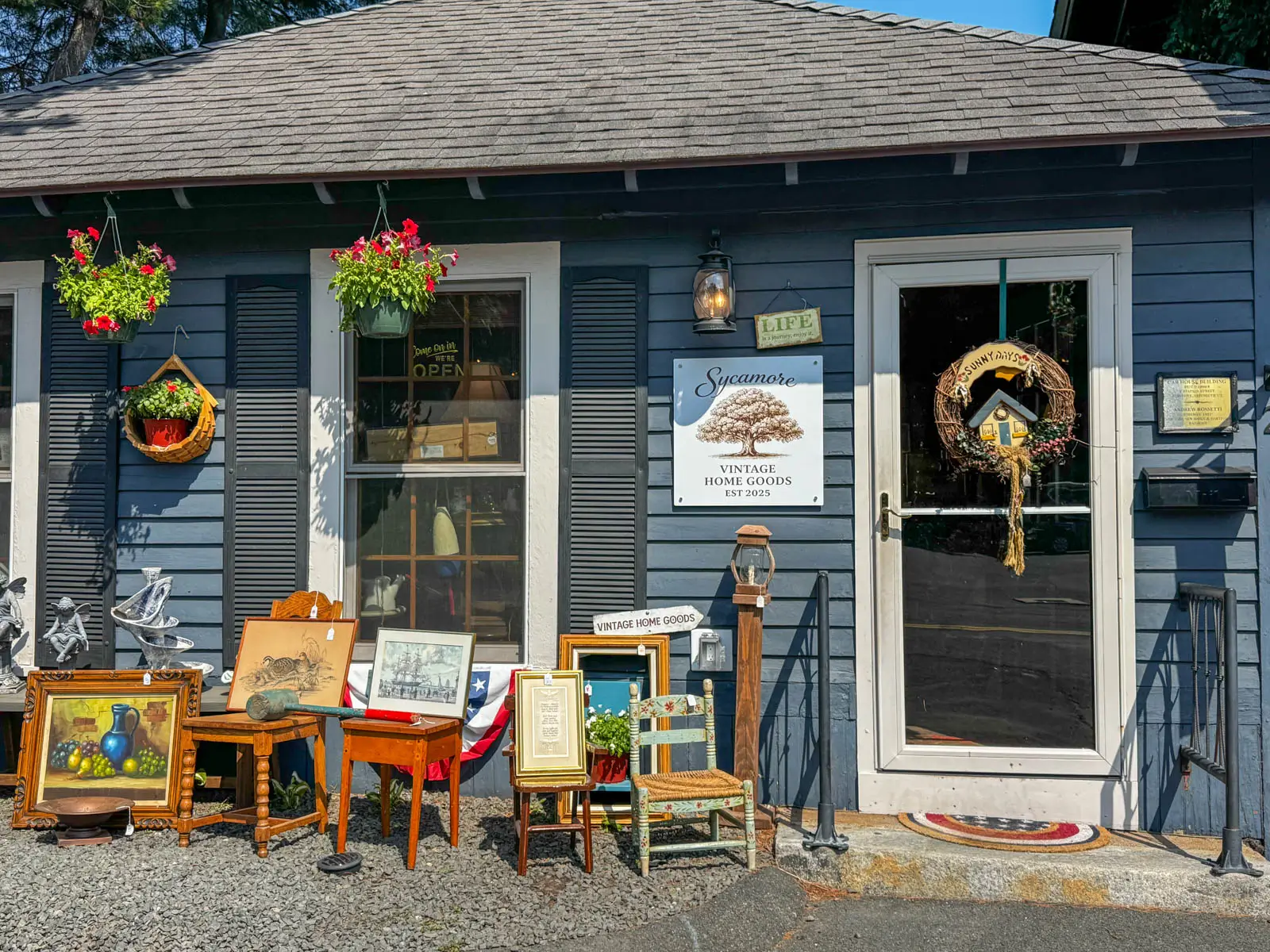 front of small blue house with "Sycamore Vintage Home Goods" white sign in front with brown writing and antiques scattered around the front of the building.