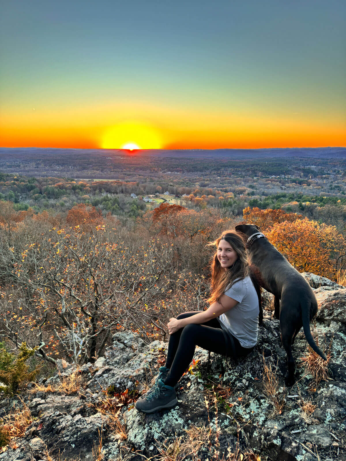 woman sitting and smiling at the top of penwood state forest hike with a sunset view of golden sun setting behind her and brown dog next to her.