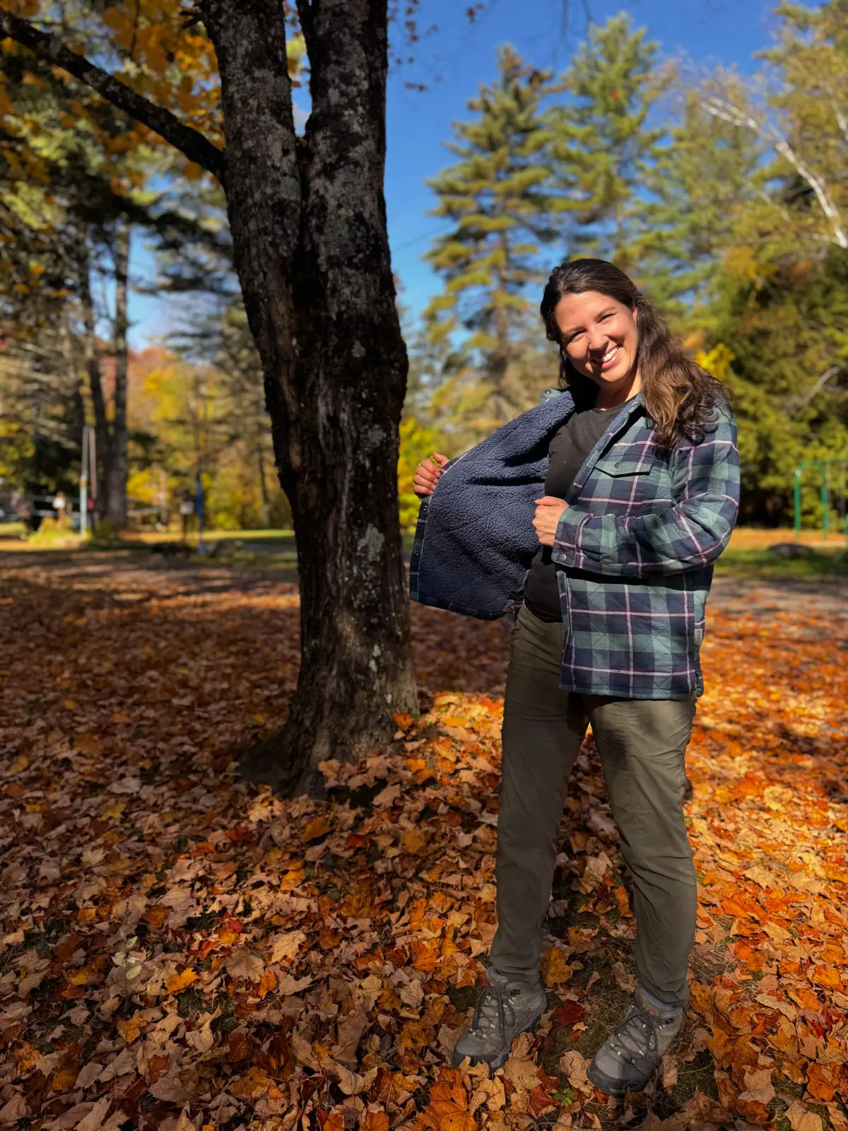 brunette woman smiling at camera standing in orange leaf pile in vermont wearing a navy blue and green checkered flannel with fleece lining on a sunny day.