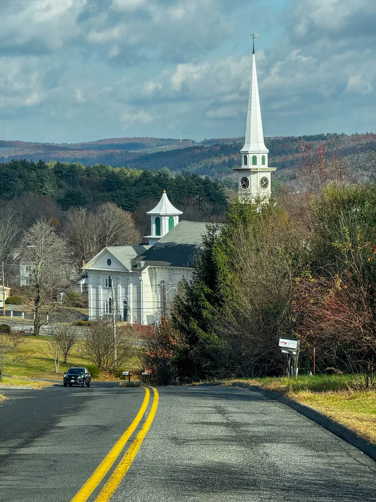 road in late fall lined with bare trees and white steeple church in distance on cloudy day in monson massachusetts.