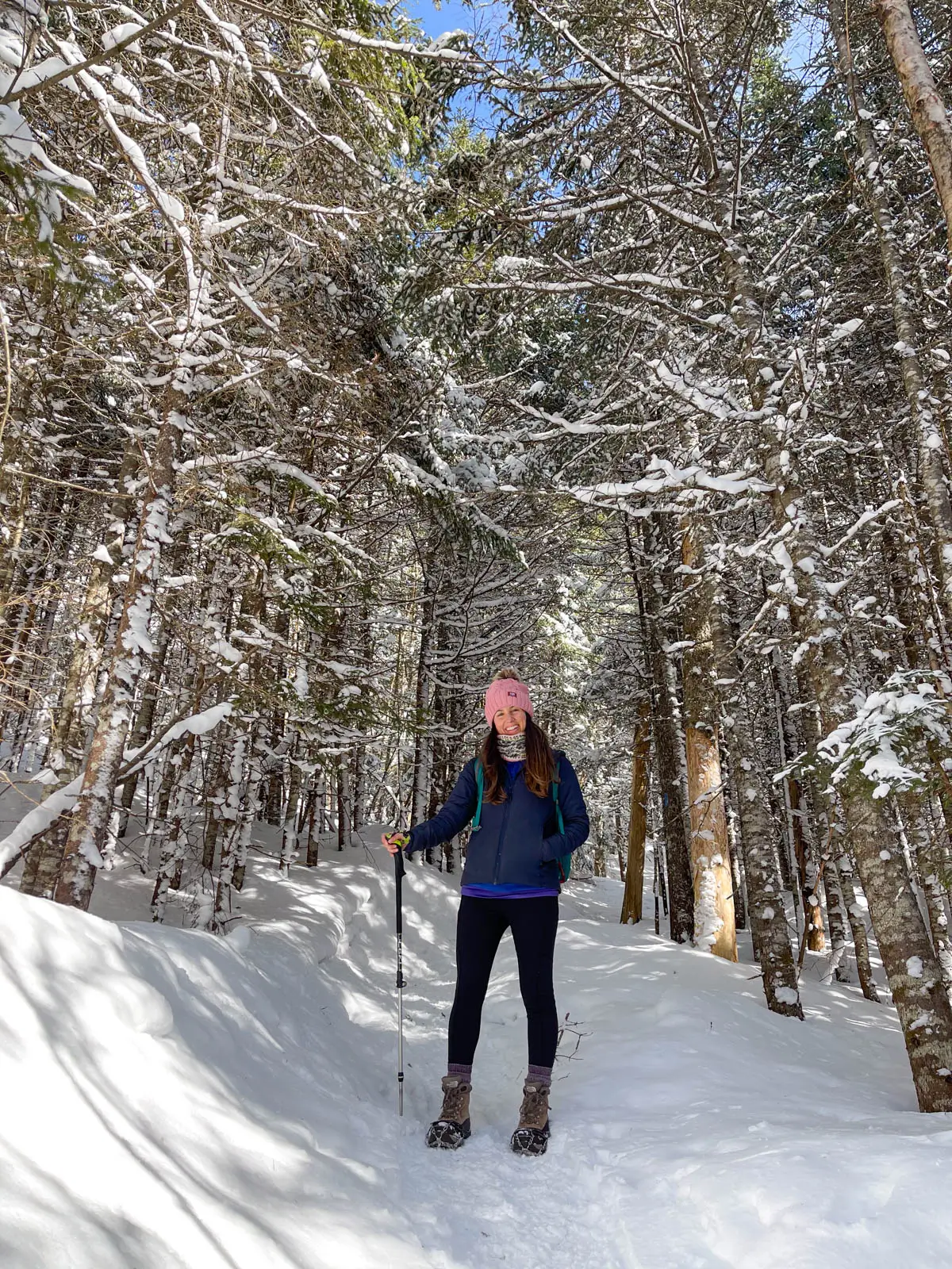 brunette woman with pink snow hat and black leggings hiking through a tunnel of snow covered trees in new hampshire.