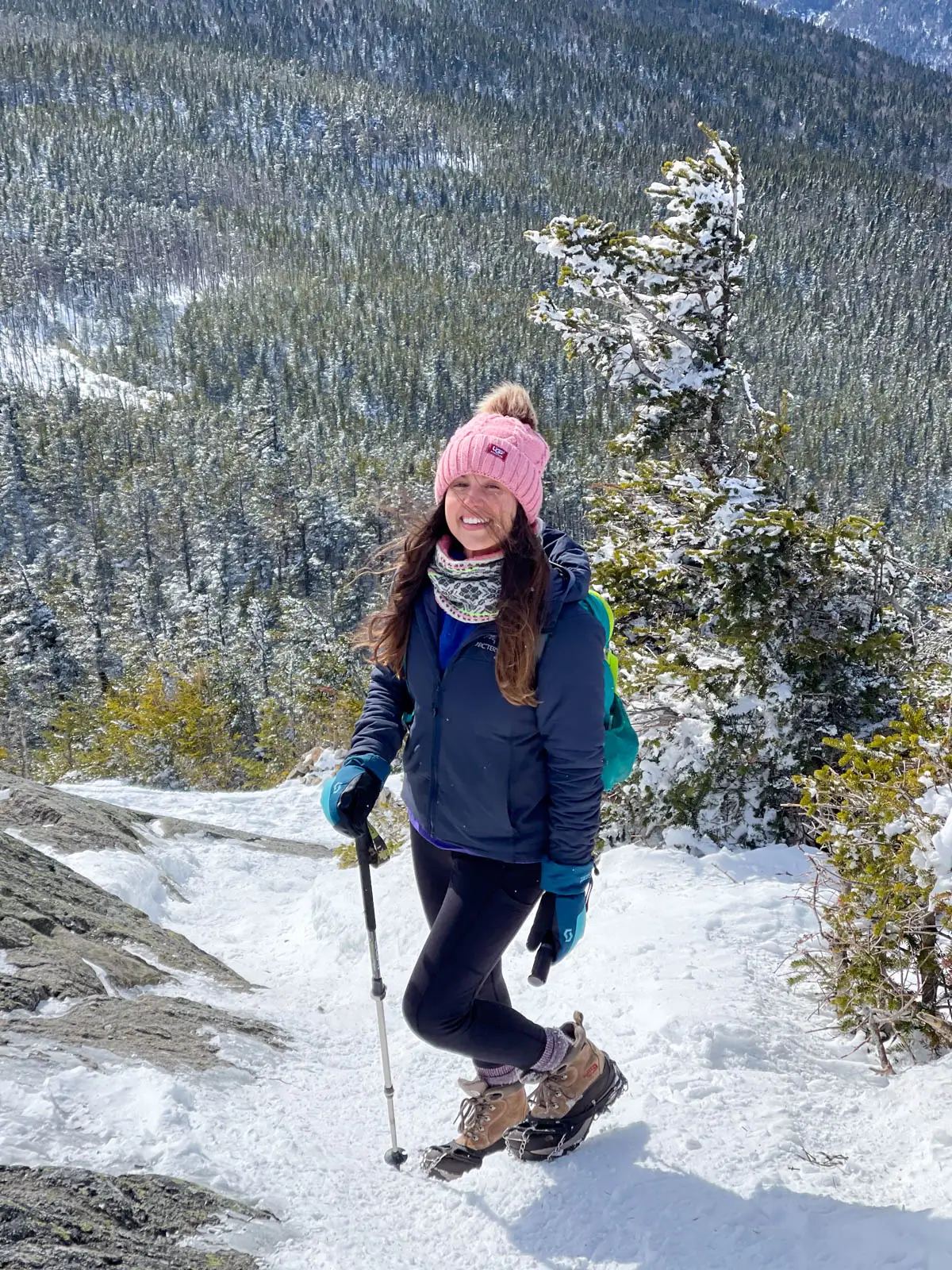 brunette woman with pink snow hat at the top of a mountain in new hampshire with black winter hiking pants on and blue skies above.
