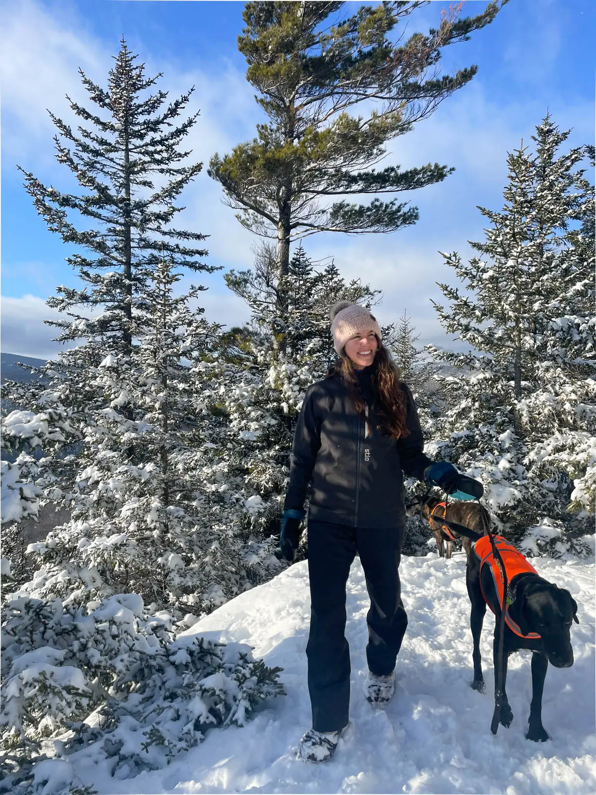 woman in black winter jacket and insulated winter pants hiking through the snow with a black dog next to her on a sunny day with blue skies.