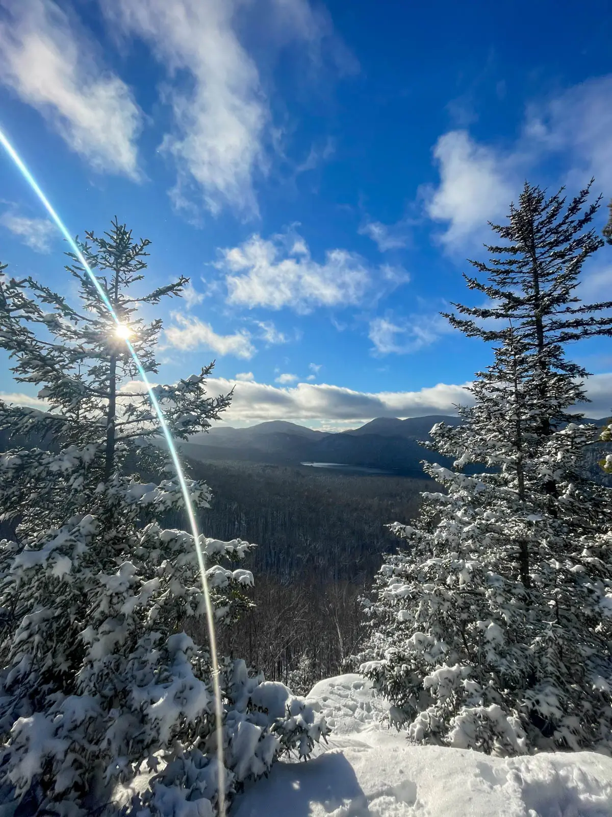 view from top of a snowy hike with two every green trees lining either side of the photo with snow on the ground and a bright sun shining in a blue sky.