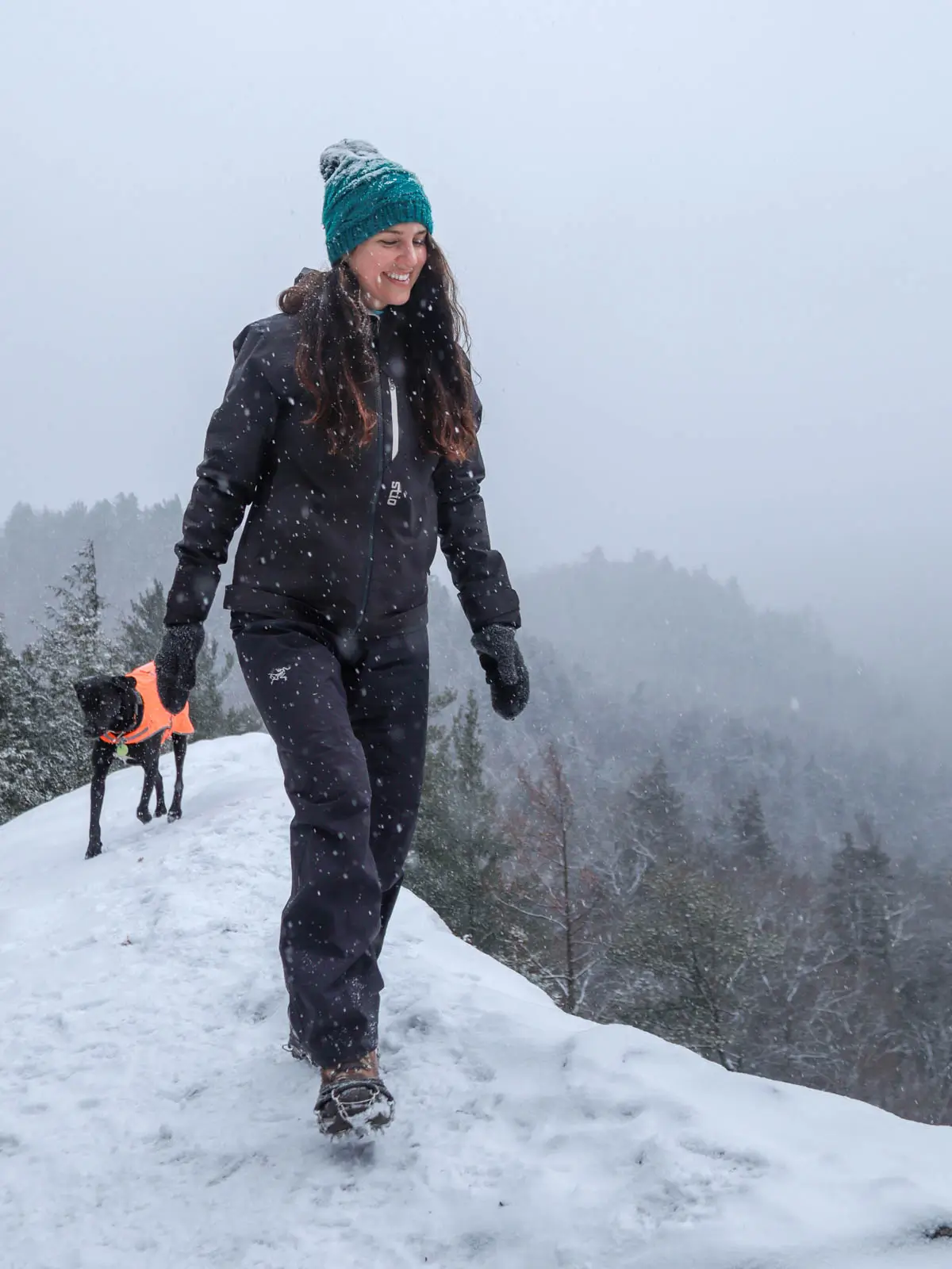 brunette woman smiling woith turquoise snow hat and black winter hiking pants and jacket walking along an edge of a snowy cliff with black dog in orange vest behind her.