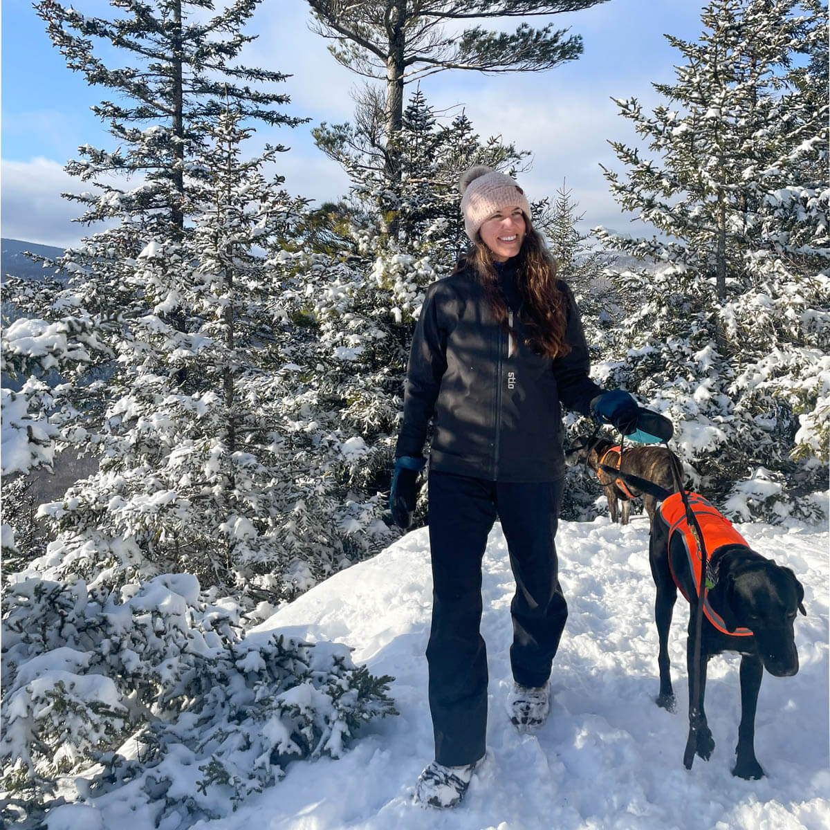 woman in black winter jacket and insulated winter pants hiking through the snow with a black dog next to her on a sunny day with blue skies.