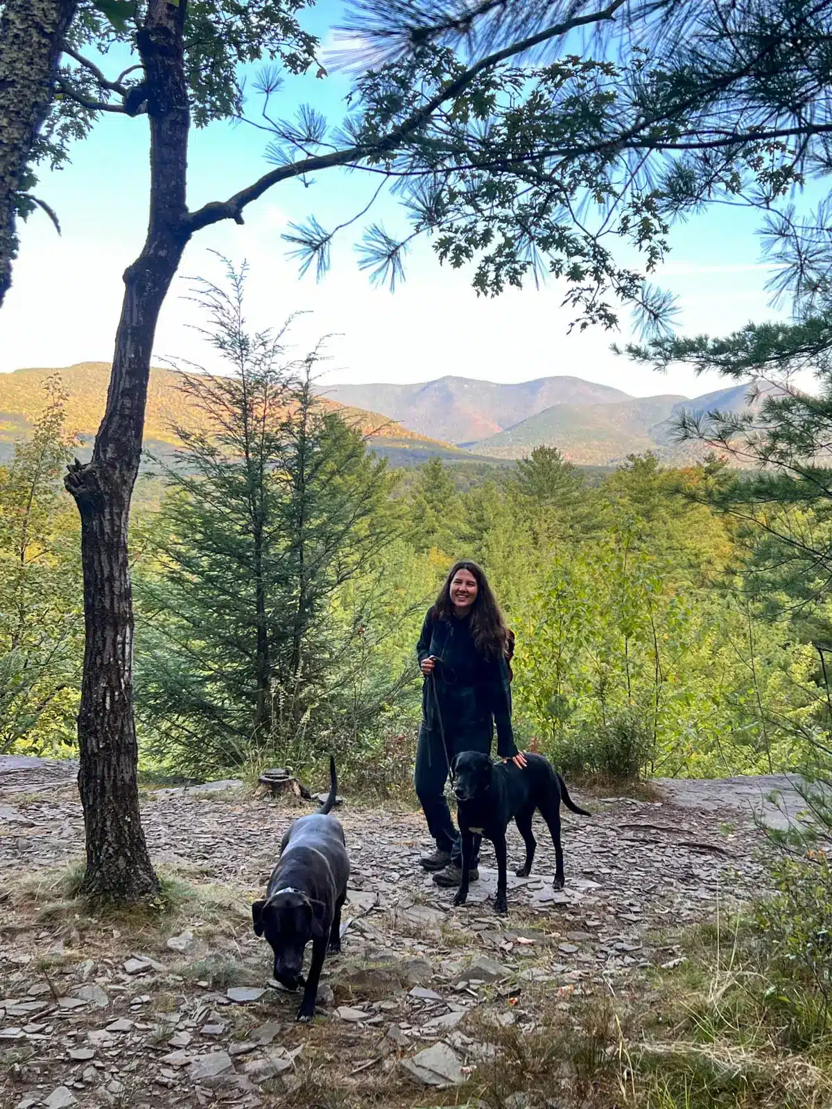 woman smiling on top of hike with green mountains and trees in background with black and brown dog at ashokan quarry.