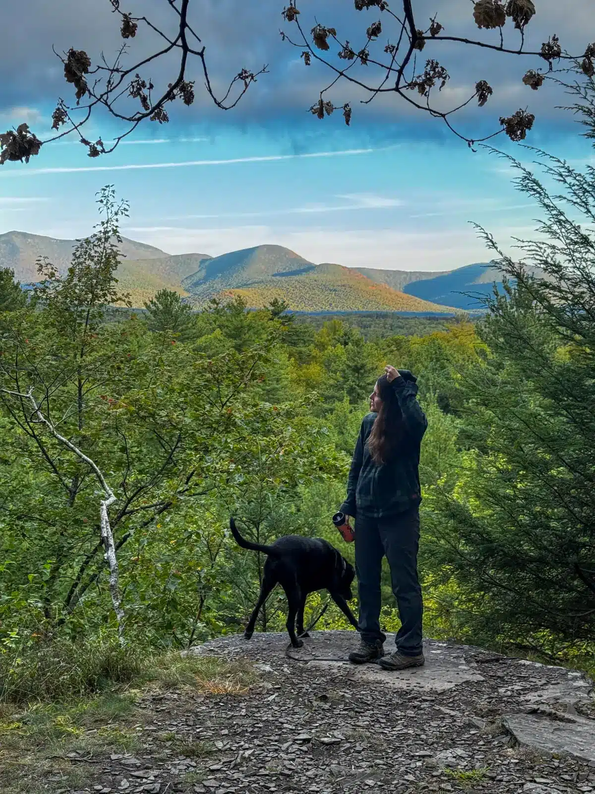 woman smiling on top of hike with green mountains and trees in background with black and brown dog at ashokan quarry.