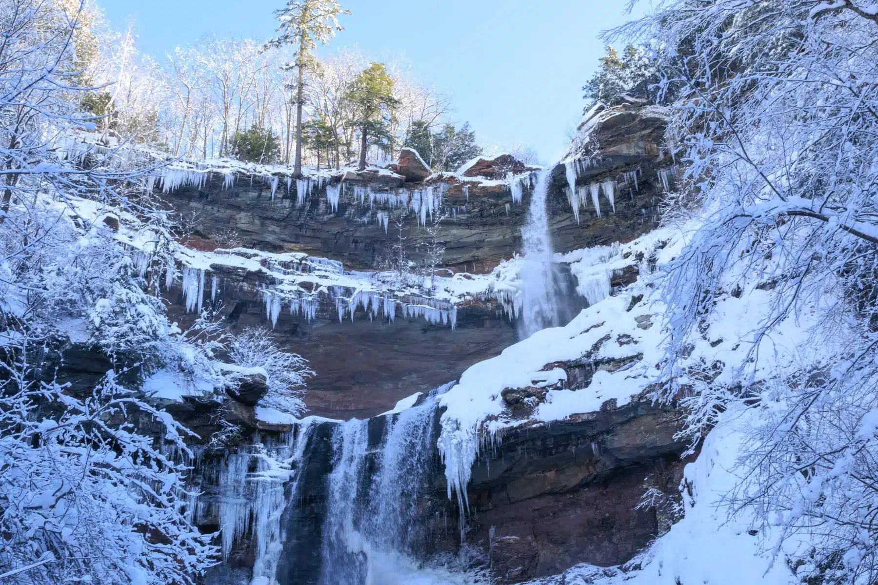kaaterskill falls two tiered frozen waterfall tumbling down surrounded by icicles on a winter day with light blue sky above.