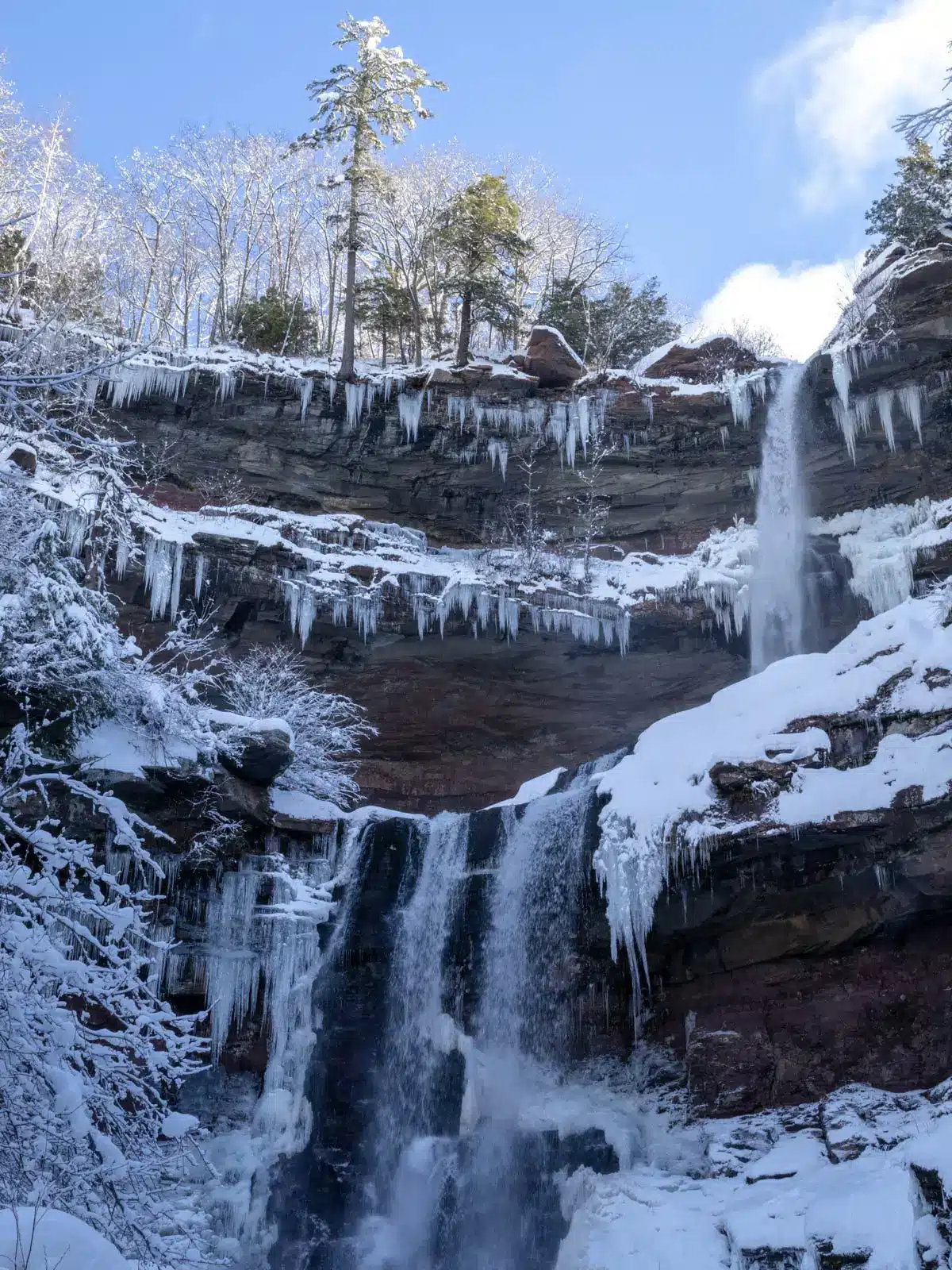 kaaterskill falls two tiered frozen waterfall tumbling down surrounded by icicles on a winter day with light blue sky above.