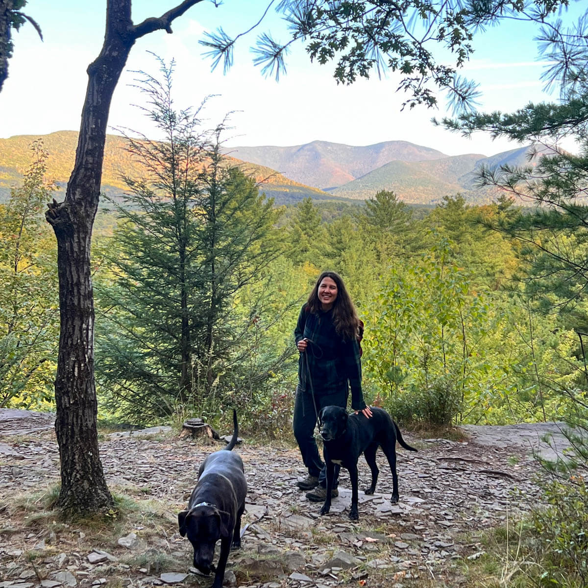 woman smiling on top of hike with green mountains and trees in background with black and brown dog at ashokan quarry.