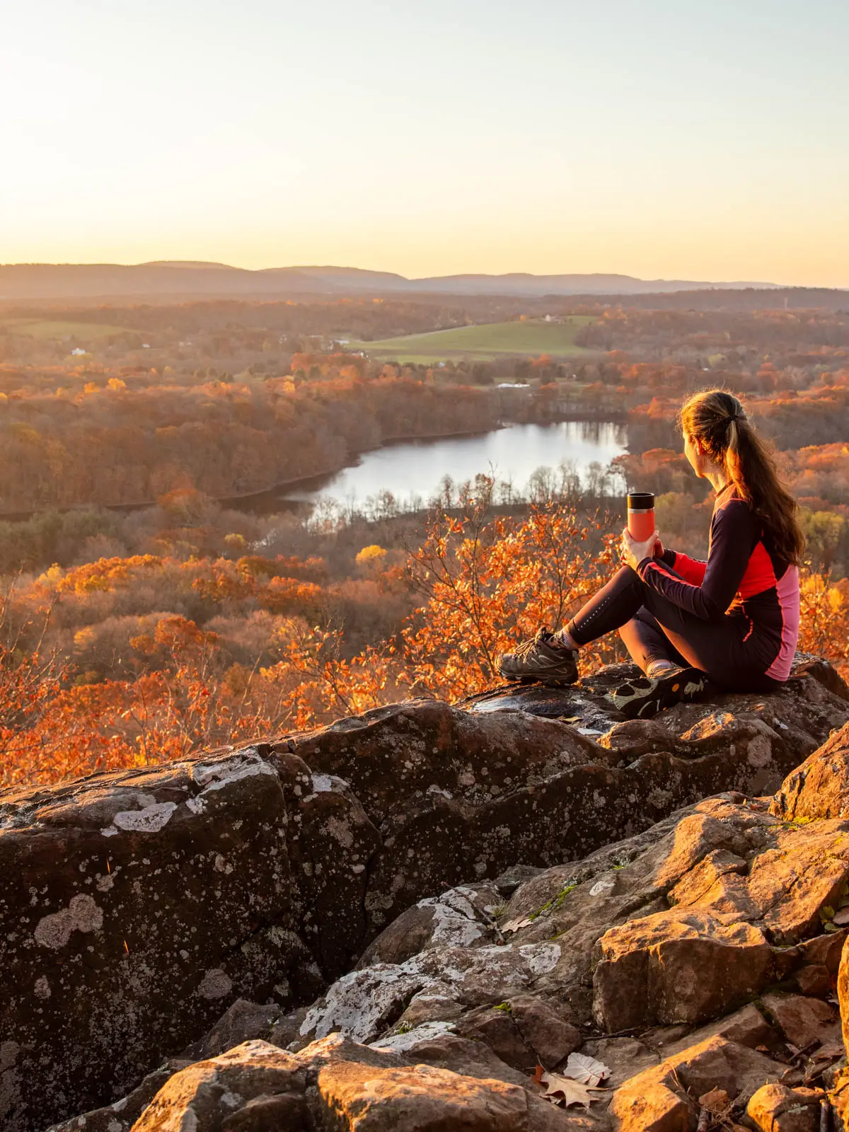 woman sitting on cliff edge looking out at hillside beauty at sunrise time with soft yellow glow in sky and orange tree tops in distance at ragged mountain.