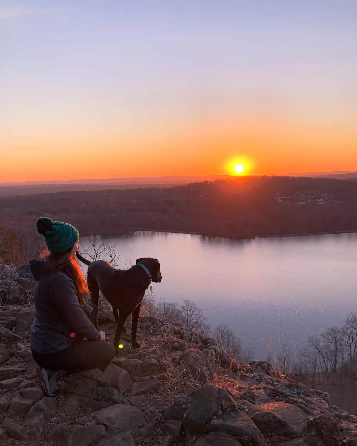 girl in snow hat and jacket kneeling beside black shiny dog at sunrise time with yellow sun coming up in distance and light blue pond below.