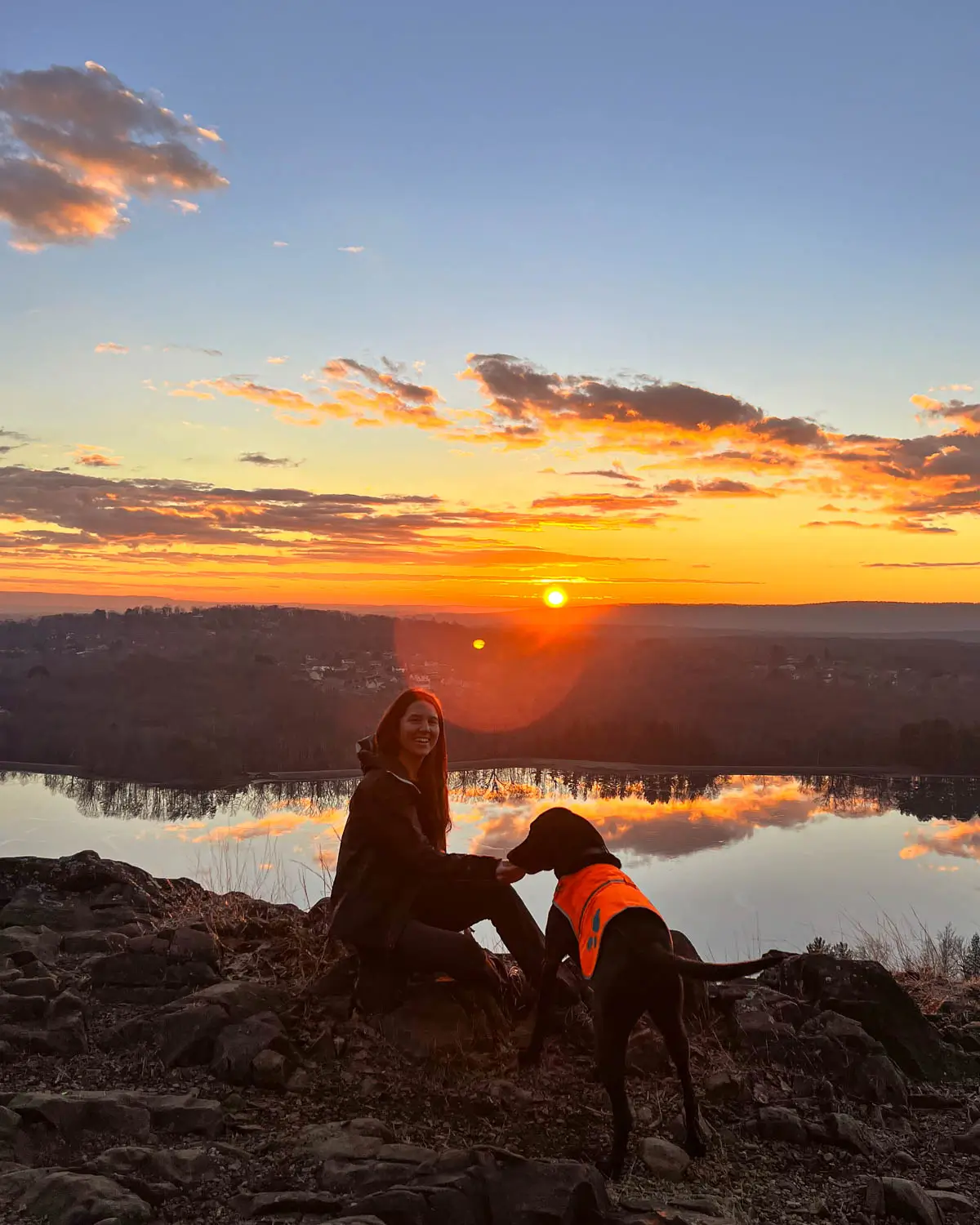 silhouette of woman and black dog looking out over light blue pond at sunrise time with yellow sun coming up over the horizon at ragged mountain in connecticut.