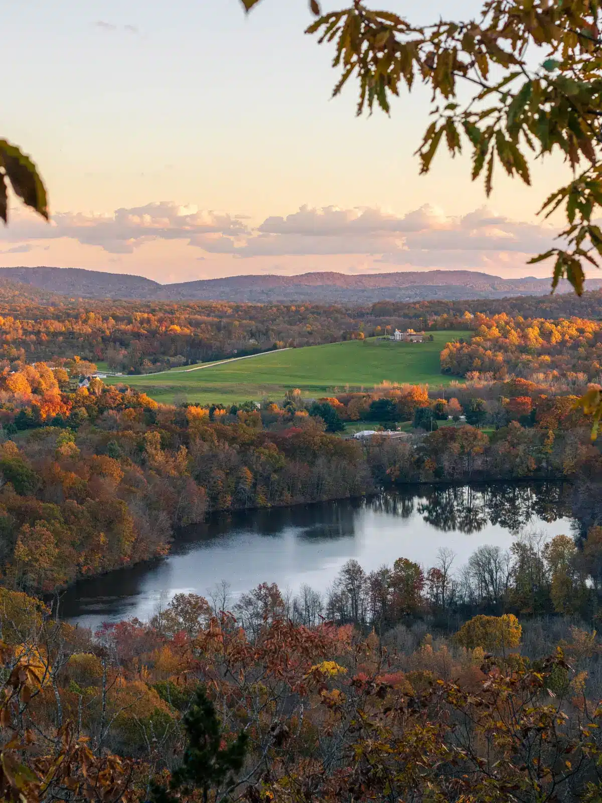 fall view from hike atop ragged mountain in Berlin with green field in distance, pond below and fall colored leaves all around.