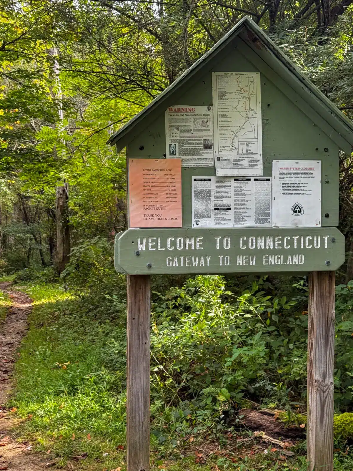 a green trailhead sign with "welcome to connecticut" on it in white letters in summer surrounded by green trees.