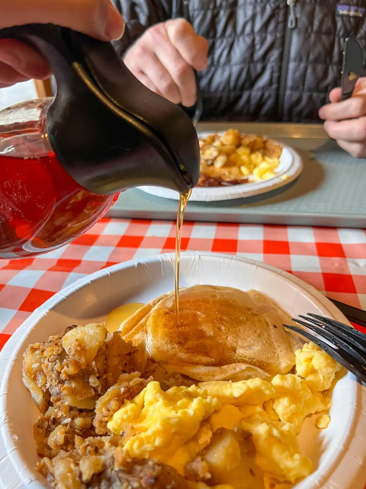 beige paper plate with pancake and eggs with maple syrup drizzled on top.