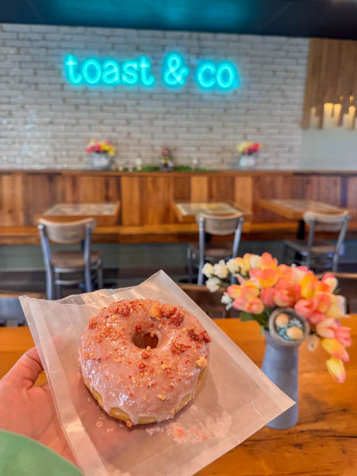 a white hand holding a light pink donut with flower vase on table and toast and co sign in background.