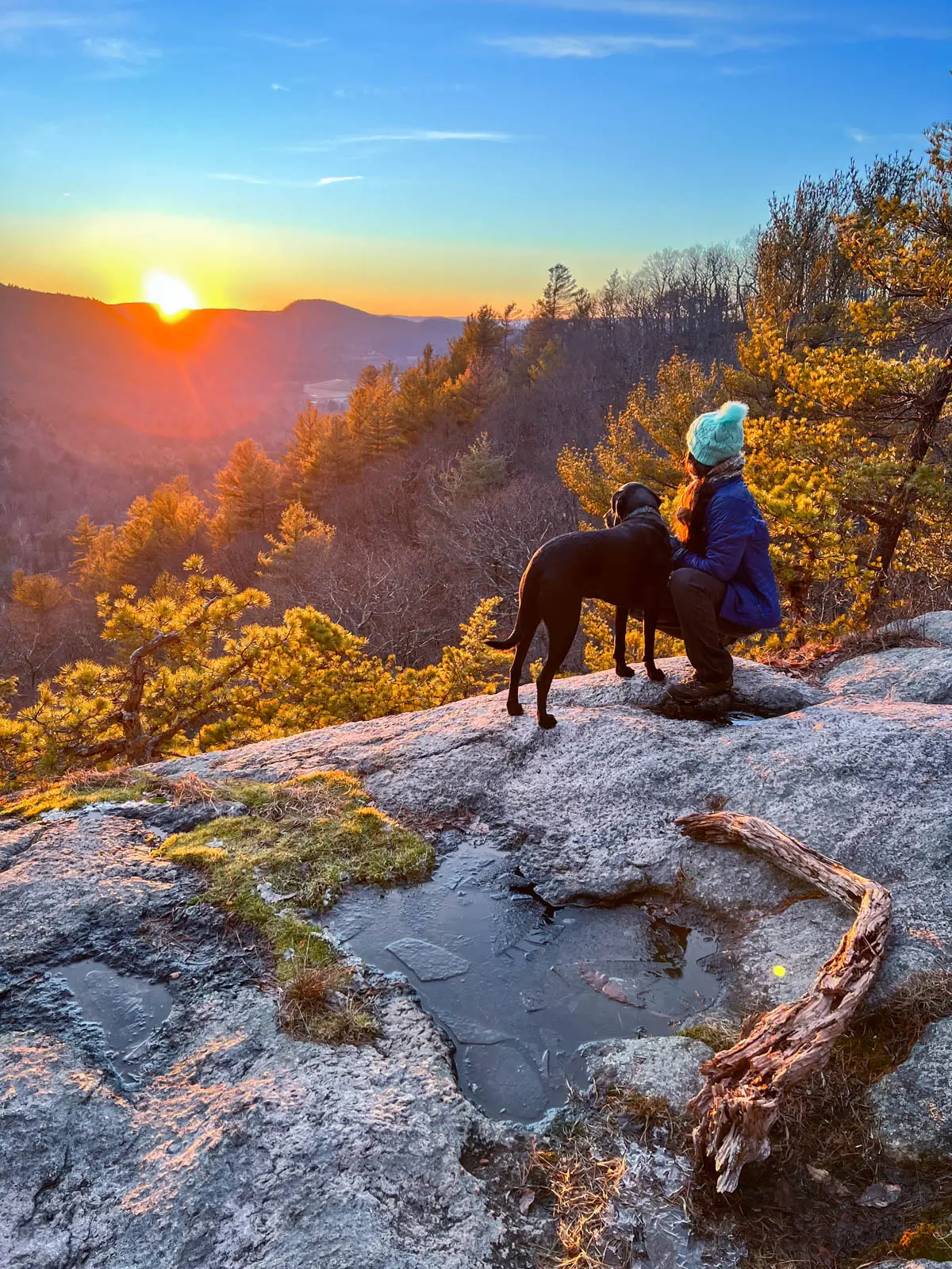 woman kneeling on mountain ledge with black dog looking out at golden sun setting at top of hike in connecticut.