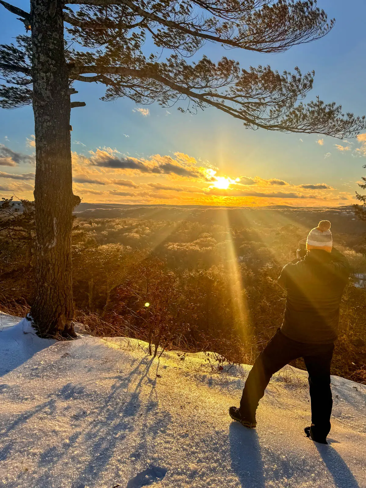 sunset view from top of hike in connecticut with golden sun setting and man in snow hat standing on cliff ledge in snow taking a photo.