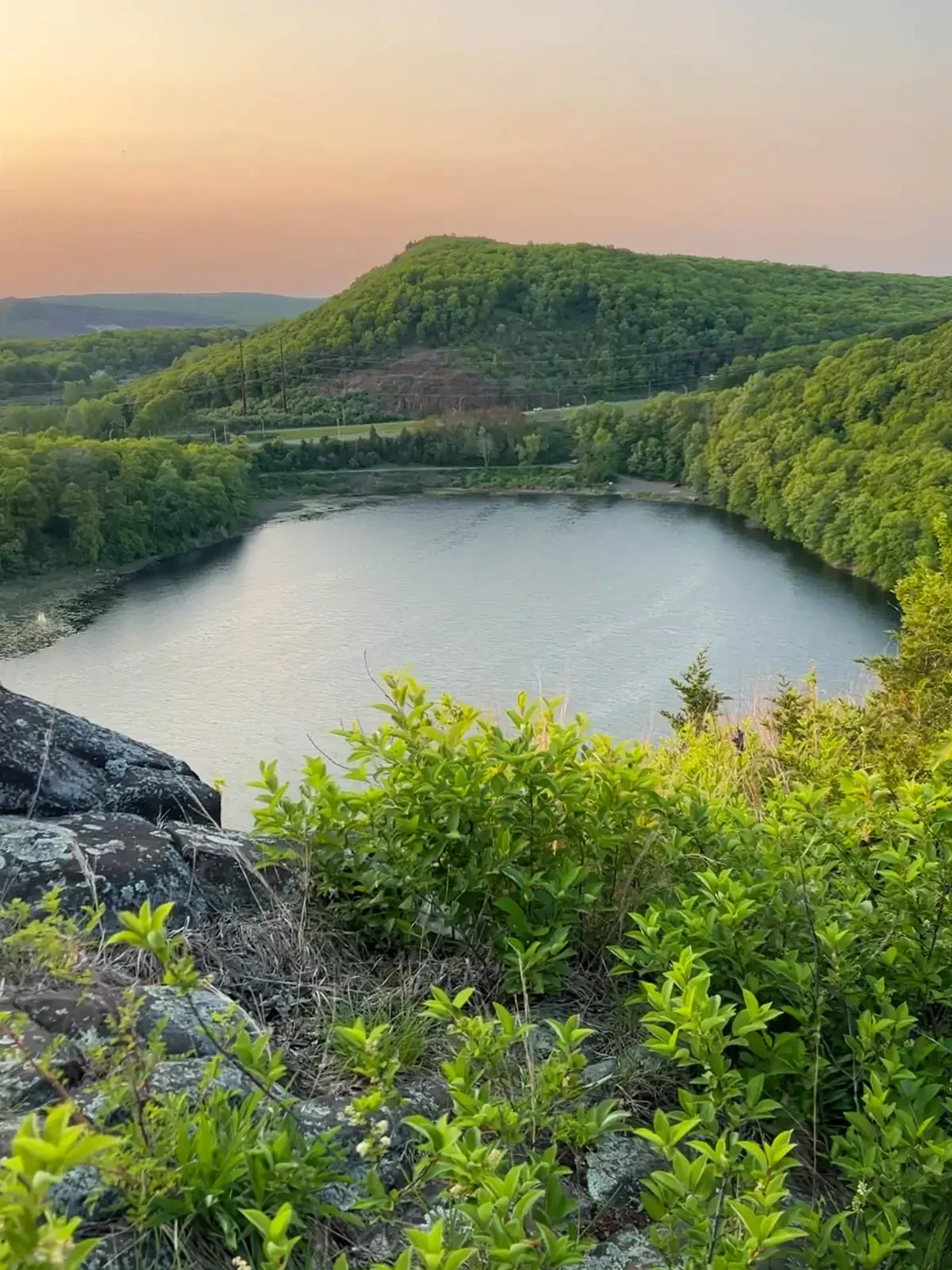 view from top of hike down at large lake surrounded by green hills and soft pastel sunset sky.