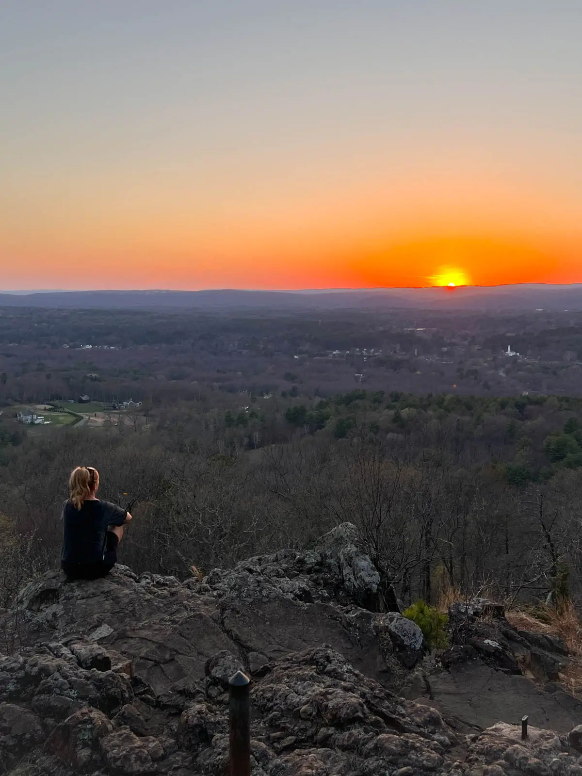 woman in black t-shirt sitting looking out from cliff on a sunset hike with yellow sun setting in distance and orange glow in sky.