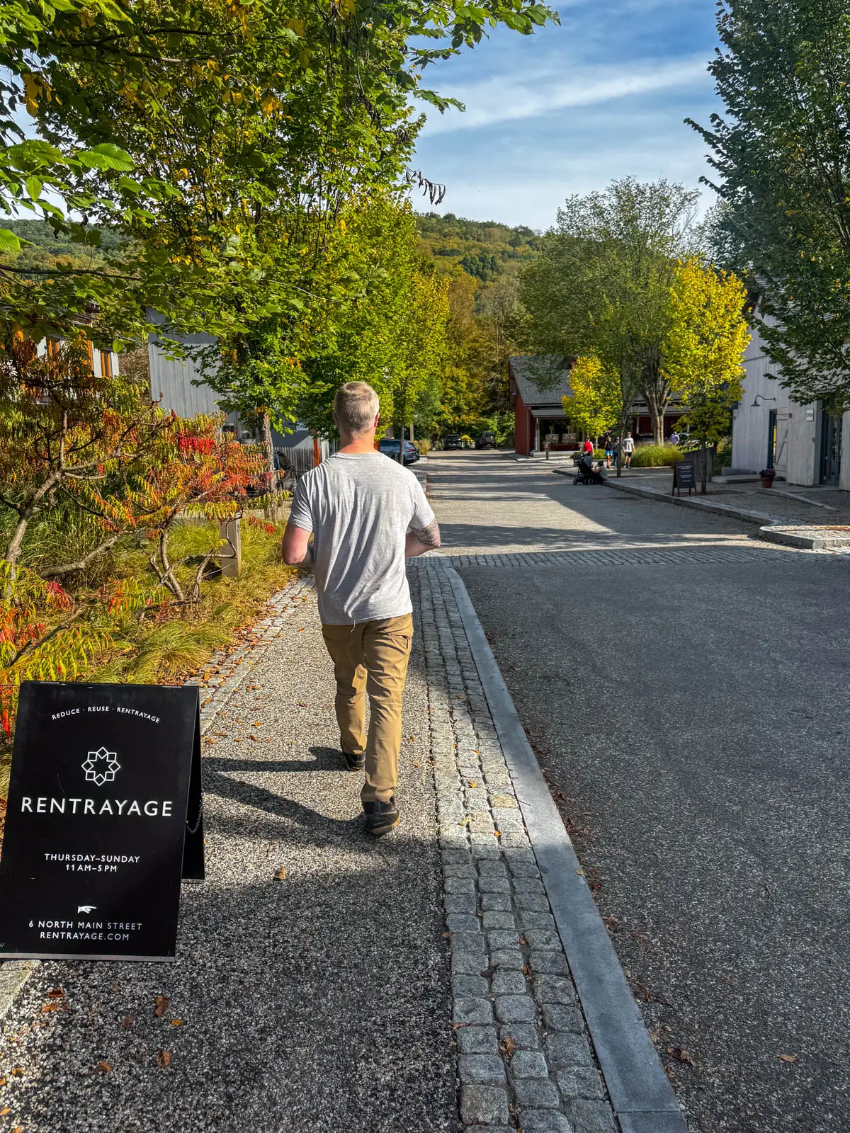back of man in gray t-shirt walking down cobblestone sidewalk with rolling green hill in distance accented with gold leaves in fall.