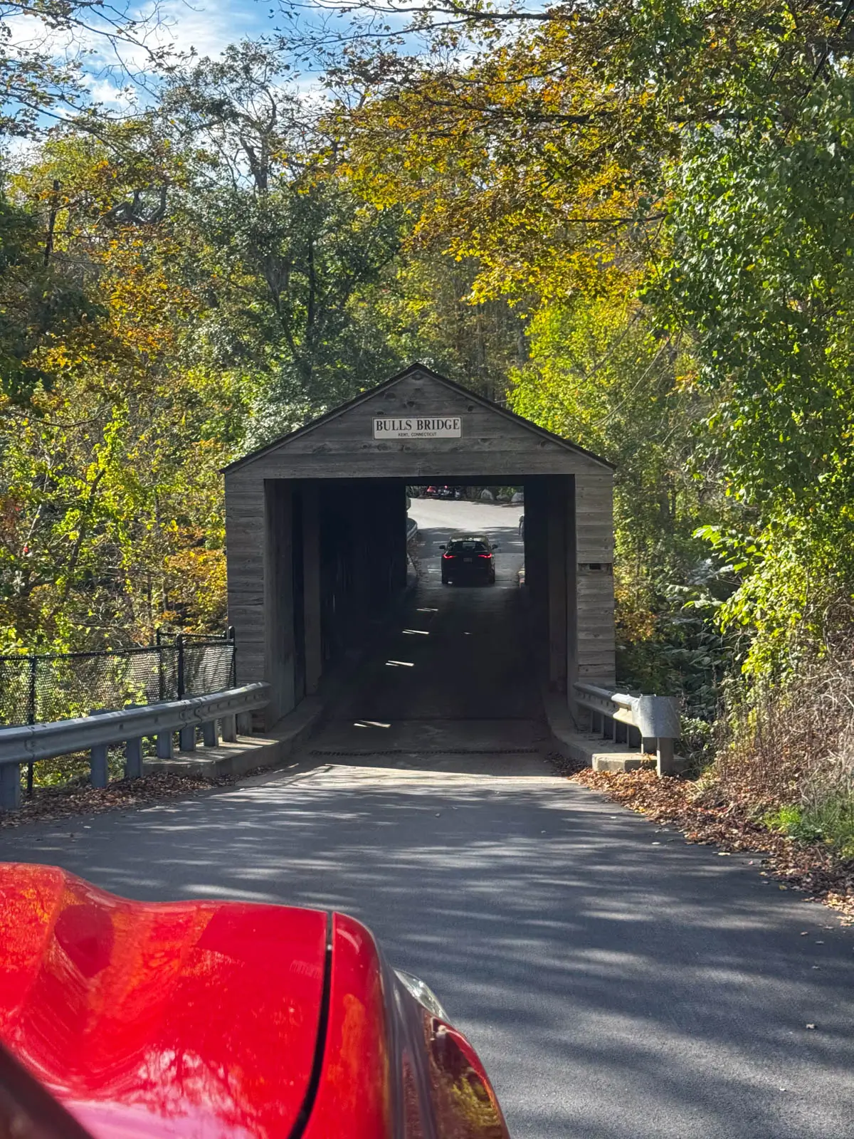 front of a bright red truck as it enters a gray covered bridge in fall with green and gold leaves framing the bridge entrance.