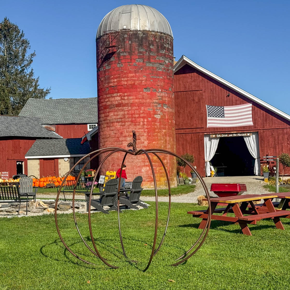 big red barn with american flag on it and silo in litchfield connecticut with blue sky above, green lawn in front, picnic tables and fall decor.