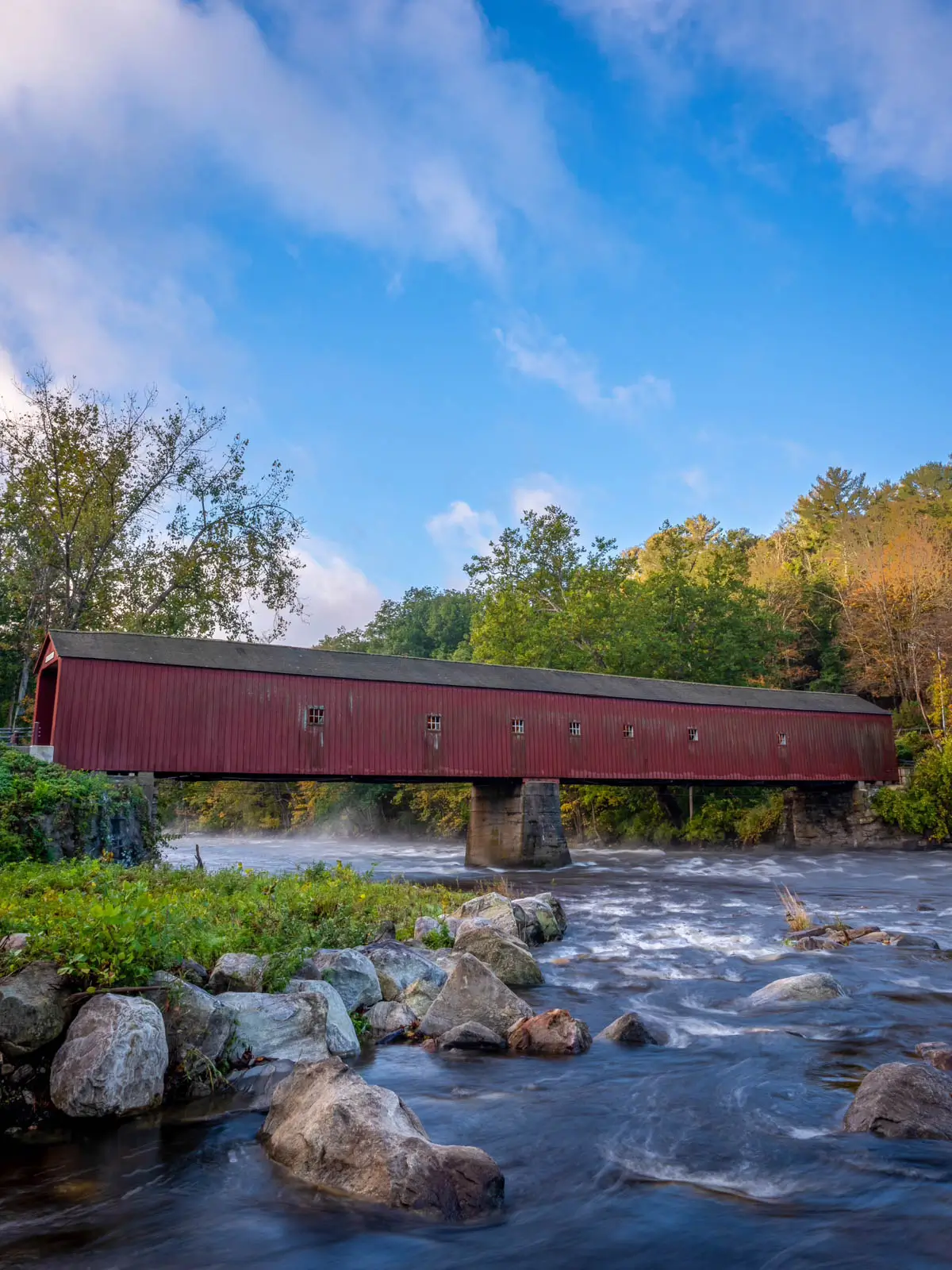 long red covered bridge in cornwall connecticut in fall surrounded by orange and yellow leaves.