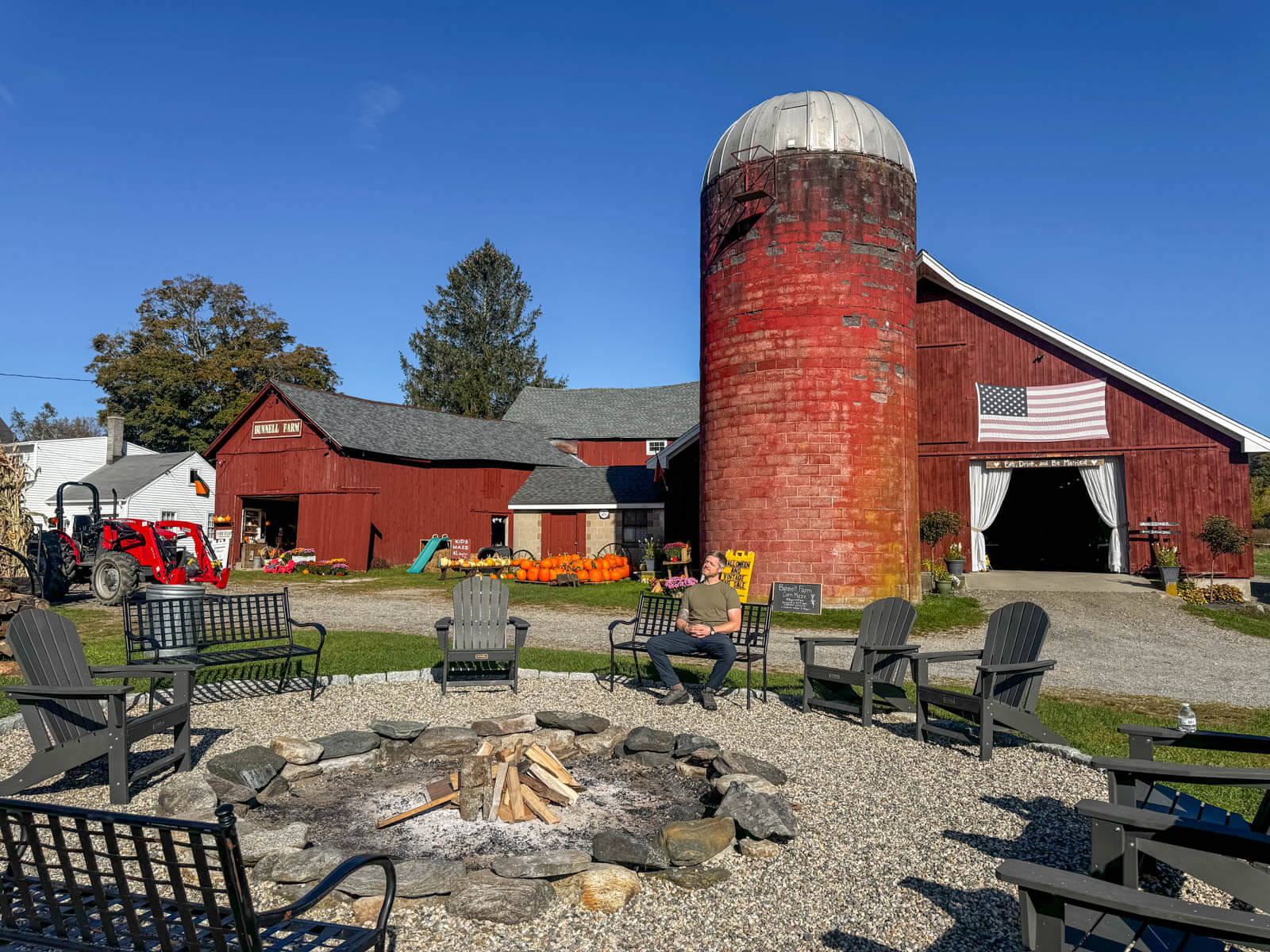 red barn with fire pit in front and adirondack chairs arranged in a circle in front with a man sitting in one of the chairs. 