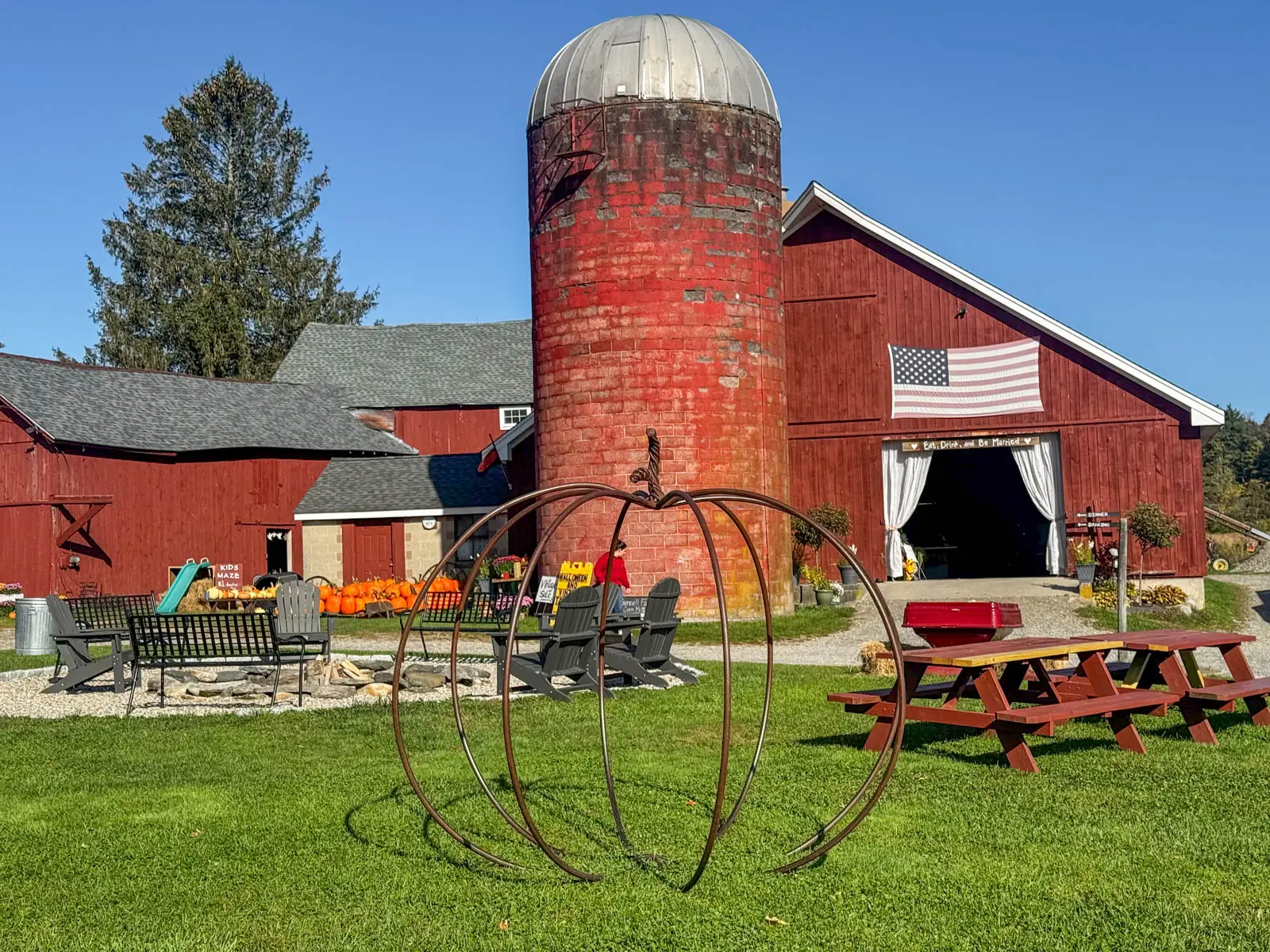 big red barn with red silo in fall with american flag on the barn and pumpkins out front.